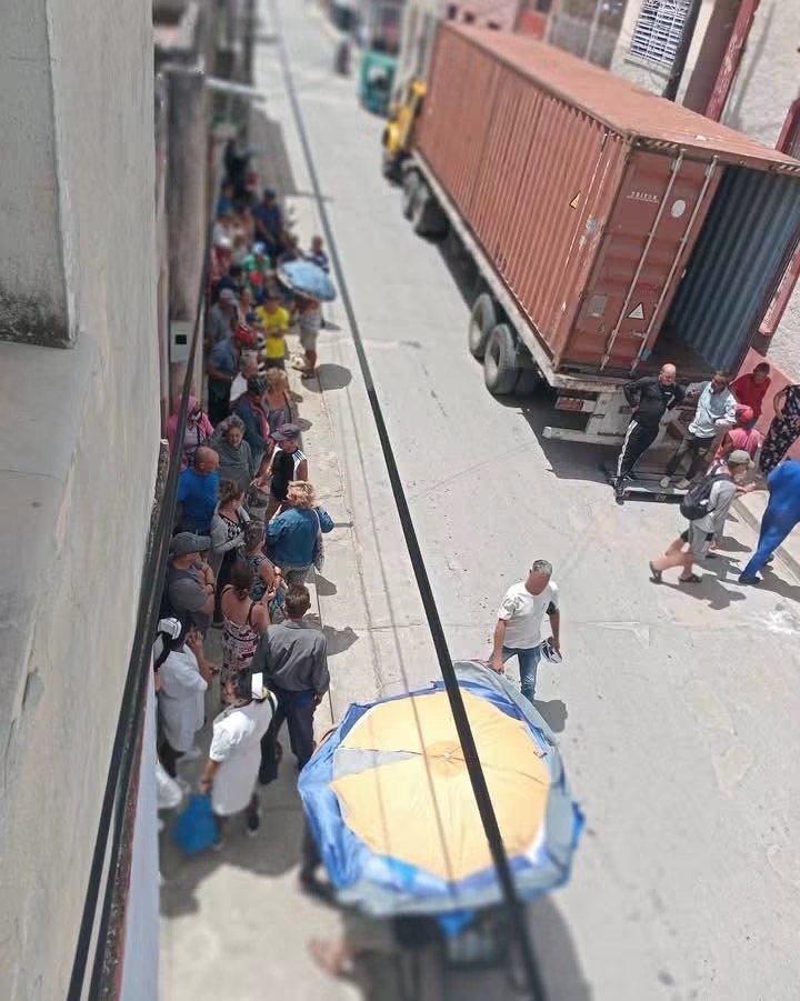 Personas hacen cola junto a una pared mientras un camión con contenedor ocupa la calle.