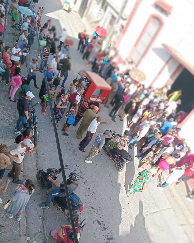 Decenas de personas esperan junto a un edificio rosado y una entrada comercial en Holguín.