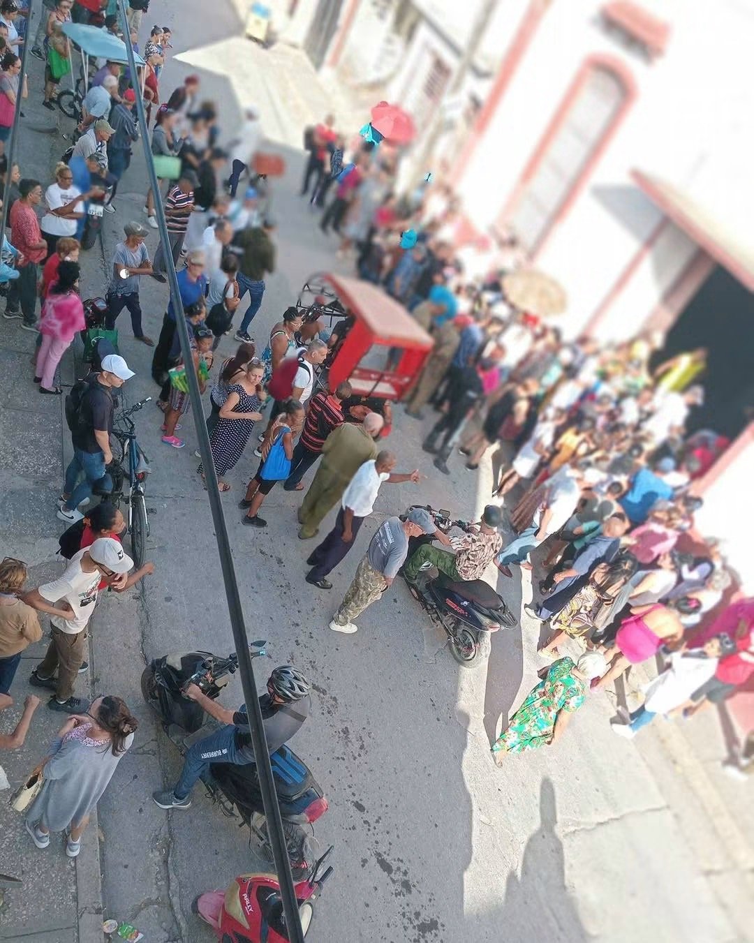 Decenas de personas esperan junto a un edificio rosado y una entrada comercial en Holguín.