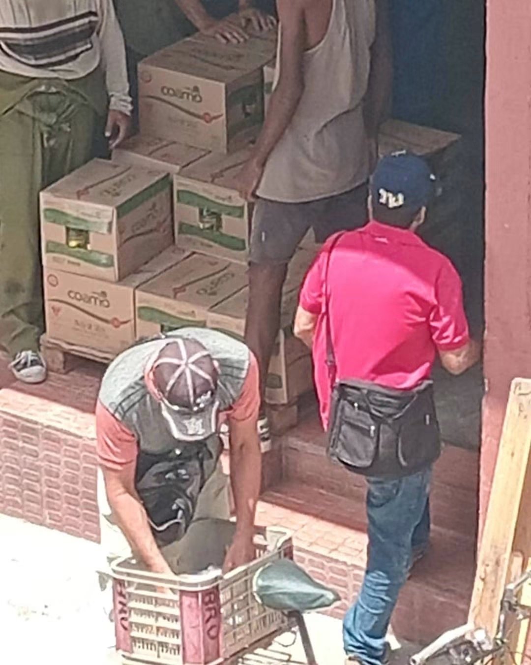 Cajas de aceite apiladas en la entrada de un establecimiento y varias personas junto a ellas.