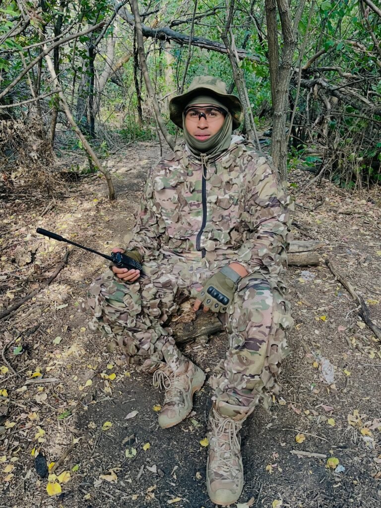 El joven conocido como El Sensei 2002, con ropa de camuflaje y una radio de mano, sentado en una zona arbolada.