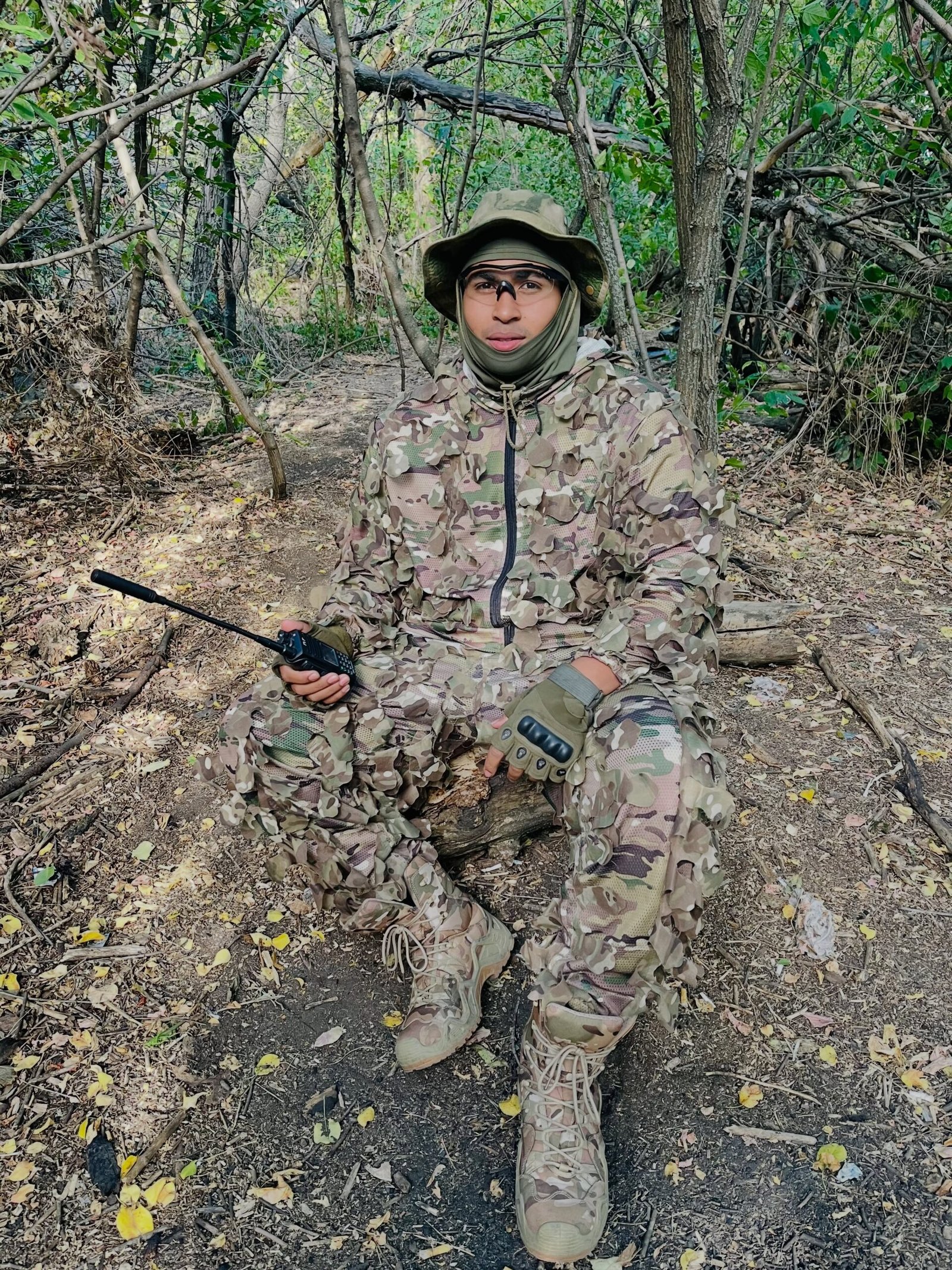 El joven conocido como El Sensei 2002, con ropa de camuflaje y una radio de mano, sentado en una zona arbolada.
